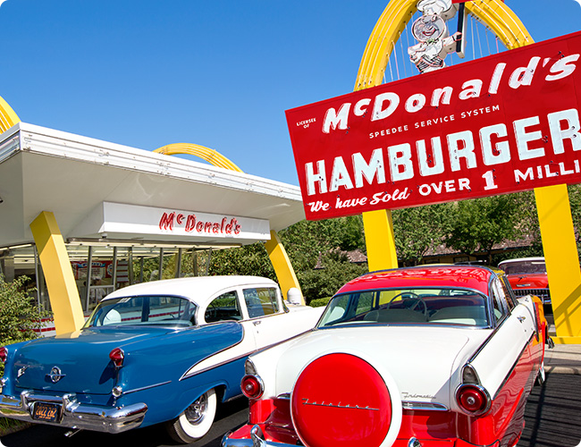 파란 하늘이 보이는 맥도날드 매장을 배경에 왼쪽에는 파란차 오른쪽에는 빨간 차가 주차되어 있는 사진