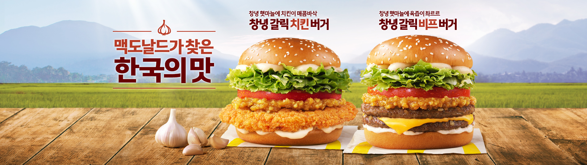 창녕 햇마늘로 만든 마늘 소스를 활용한 맥도날드 한국의 맛 한정 메뉴 소개 배너 이미지. 왼쪽에는 &#39;창녕 갈릭 치킨버거&#39;, 오른쪽에는 &#39;창녕 갈릭 비프버거&#39;가 나란히 진열되어 있으며, 배경은 논과 산이 펼쳐진 한국 시골 풍경. 중앙에는 &#39;맥도날드가 찾은 한국의 맛&#39;이라는 문구가 강조되어 있음.