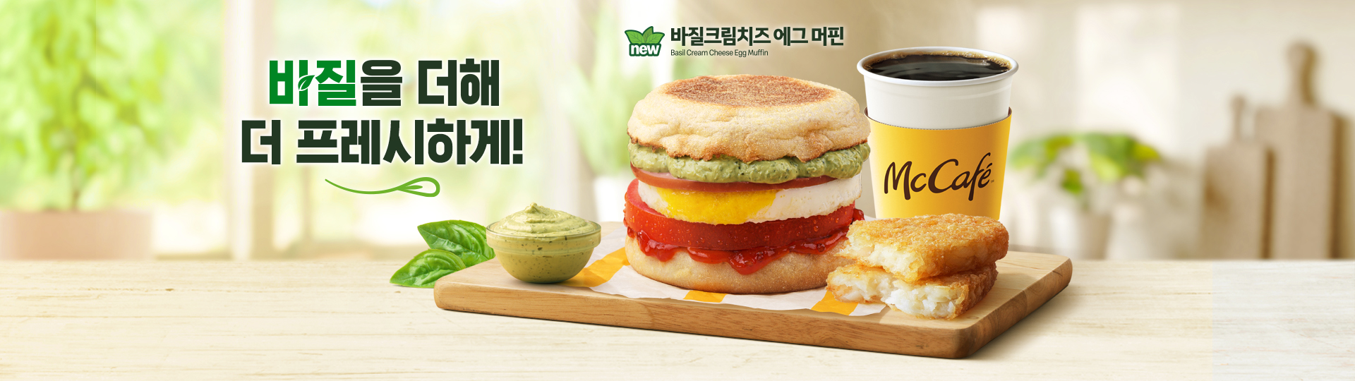 전체적으로 밝고 산뜻한 톤의 배경과 바질 잎 요소를 활용해 신선하고 프레시한 이미지이며,  “바질을 더해 프레시한 아침”라는 문구가 상단에 크게 배치되어 있다. 중앙에는 잉글리시 머핀 번 사이에 계란, 토마토, 바질 크림 소스가 들어간 ‘바질 크림치즈 에그 머핀’이 메인으로 놓여 있으며, 제품 아래에는 종이 포장지가 깔려 있다. 우측에는 따뜻한 커피가 함께 배치되어 있고, 전면 우측에는 바삭하게 구워진 해시브라운이 반으로 나뉘어 속이 보이도록 놓여 있다. 좌측 전면에는 바질 크림 소스가 담긴 소스 컵과 신선한 바질 잎이 함께 배치되어 제품의 신선함을 강조한다.