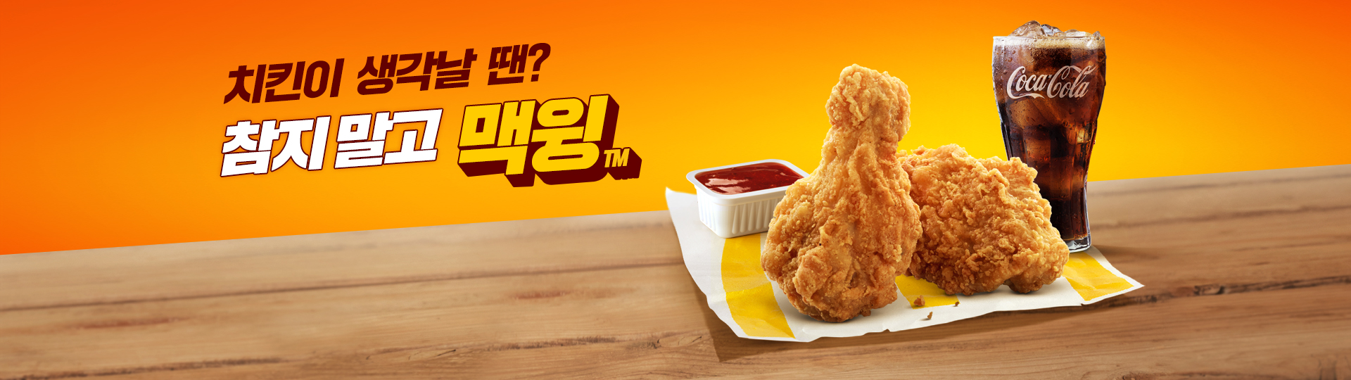 노란색 그라데이션 배경 위에 나무 테이블이 놓여 있고, 테이블 위에는 치킨 윙 두 조각이 노란 종이 포장지 위에 놓여 있으며, 옆에는 사각 흰색 소스 용기에 붉은색 소스가 담겨 있으며, 오른쪽에는 얼음이 들어간 콜라가 함께 배치되어 있다. 좌측 상단에는 ‘겉은 바삭! 속은 촉촉!’ 문구와 함께 new 배지가 붙은 겉바속촉 맥윙 제품명이 크게 노출된 맥도날드 배너 이미지