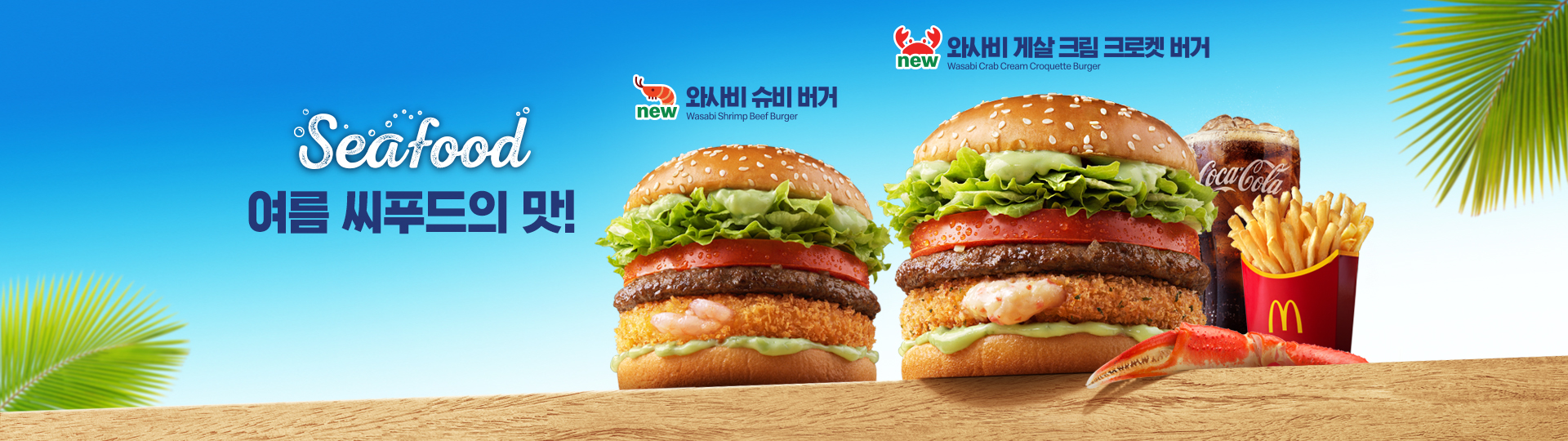 밝은 파란색 바다를 연상시키는 하늘 배경과 야자수 잎 장식 배경, 나무 테이블 위에 맥도날드 신메뉴인 와사비 슈비 버거와 와사비 게살 크림 크로켓 버거 2종이 나란히 놓여 있다.  와사비 슈비 버거에는 참깨 번 사이에 새우 패티와 비프패티, 토마토, 양상추, 와사비 타르타르 소스가 들어가있으며, 와사비 게살 크림 크로켓 버거에는 참깨 번 사이에 게살크림크로켓과 비프패티, 토마토, 양상추, 와사비 타르타르 소스가 들어가있다. 두 버거의 좌측에는 '씨푸드 여름의 맛' 문구가 있고, 와사비 게살 크림 크로켓 우측에는 맥도날드 감자튀김과 코카-콜라 음료, 집게 다리가 함께 배치되어 있으며, 전반적으로 여름 시즌 씨푸드 버거콘셉트를 강조한 광고 이미지다.