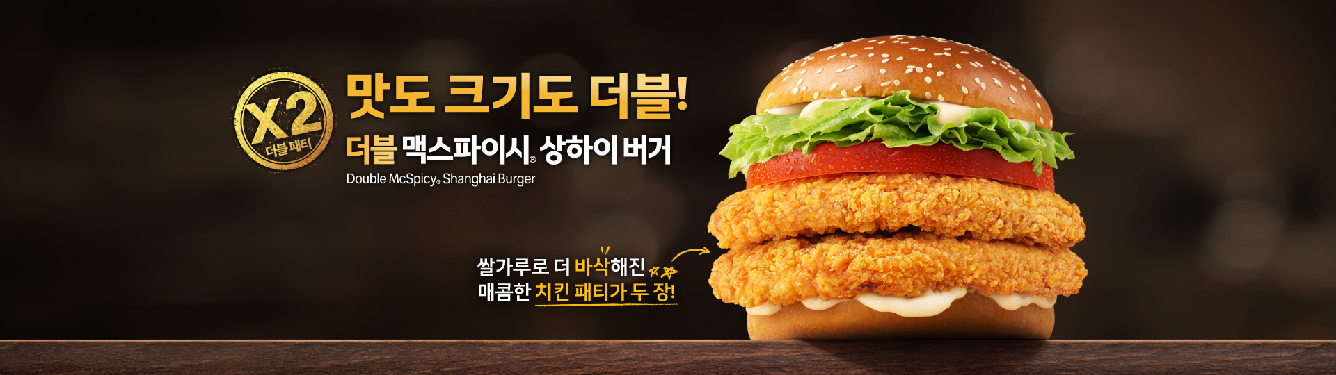 더블 맥스파이시 상하이 버거 제품 이미지. 참깨 번 사이에 양상추, 토마토, 매콤한 치킨 패티 두 장과 소스가 들어 있으며, 맛도 크기도 두 배라는 메시지를 강조하고 있다.