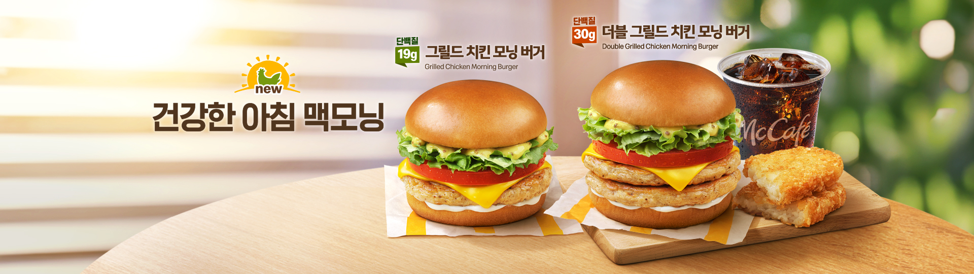 맥도날드 맥모닝 신제품을 홍보하는 가로형 메인 배너.  따뜻한 아침 햇살과 초록빛 배경을 바탕으로, 나무 테이블 위에 그릴드 치킨 모닝 버거와 더블 그릴드 치킨 모닝 버거가 놓여 있다.  왼쪽에 있는 ‘그릴드 치킨 모닝 버거’위에는 제품명과 단백질 19g이 표시되어 있다. 오른쪽에 있는 ‘더블 그릴드 치킨 모닝 버거’ 위에는 제품명과 단백질 30g이 표시되어 있고 그 옆에  아이스 커피와 해시브라운이 함께 놓여 있다. 그릴드 치킨 모닝 버거의 좌측에는 ‘건강한 아침 맥모닝’이라는 메인 카피가 쓰여져 있다.